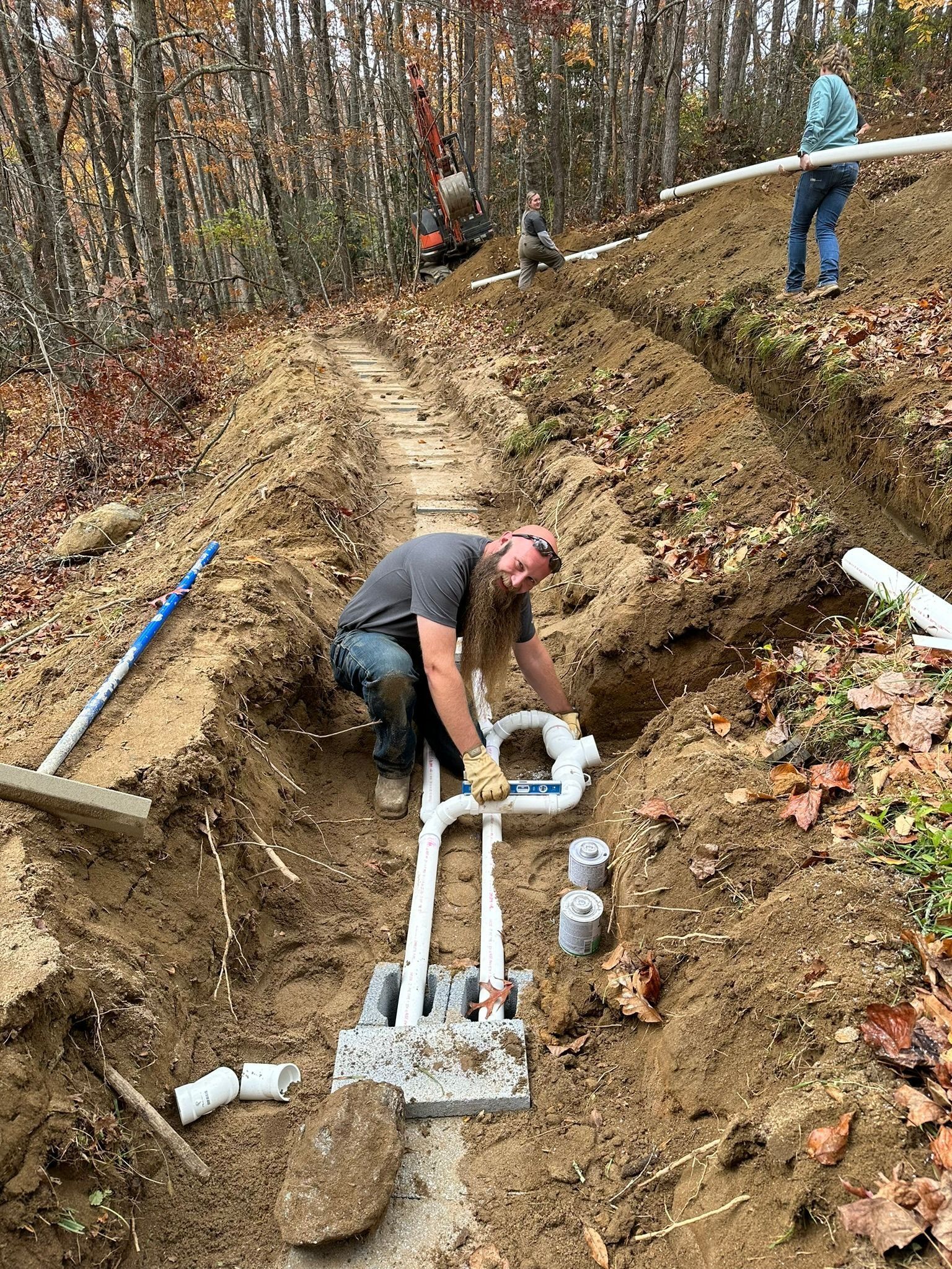 Workers installing plumbing in a trench outdoors, near trees. One man connects pipes, others work nearby.