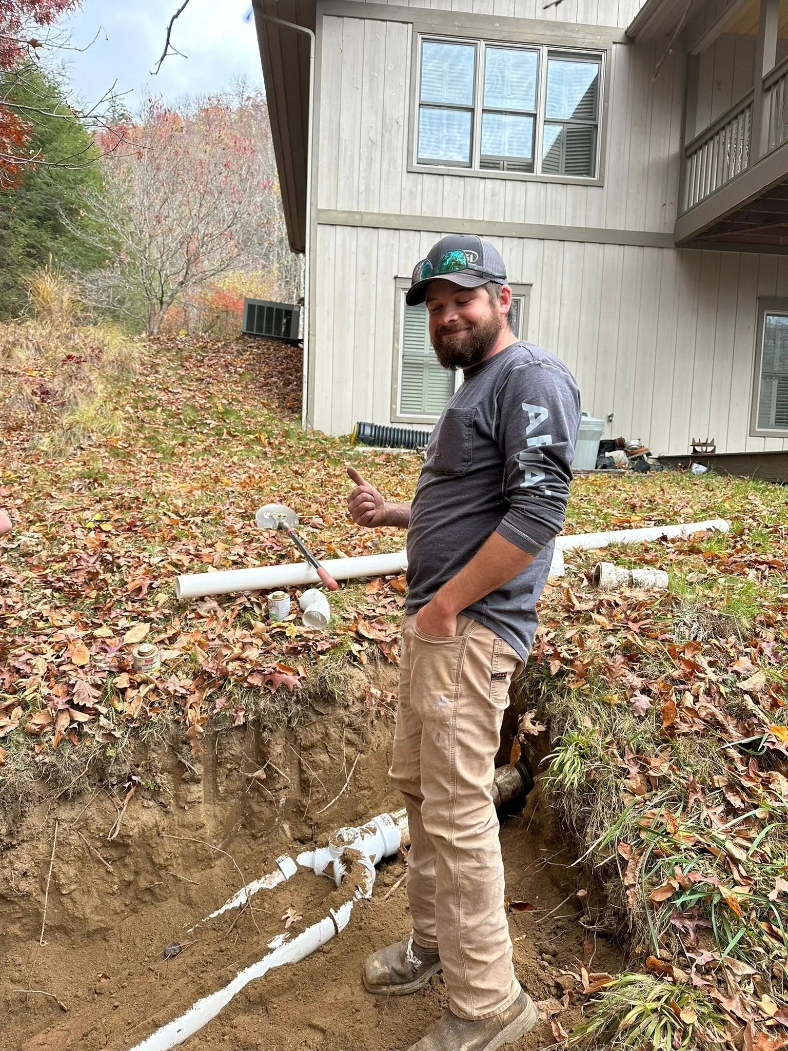 Man in work clothes beside an open trench with white pipes, near a house with windows; outdoor setting.
