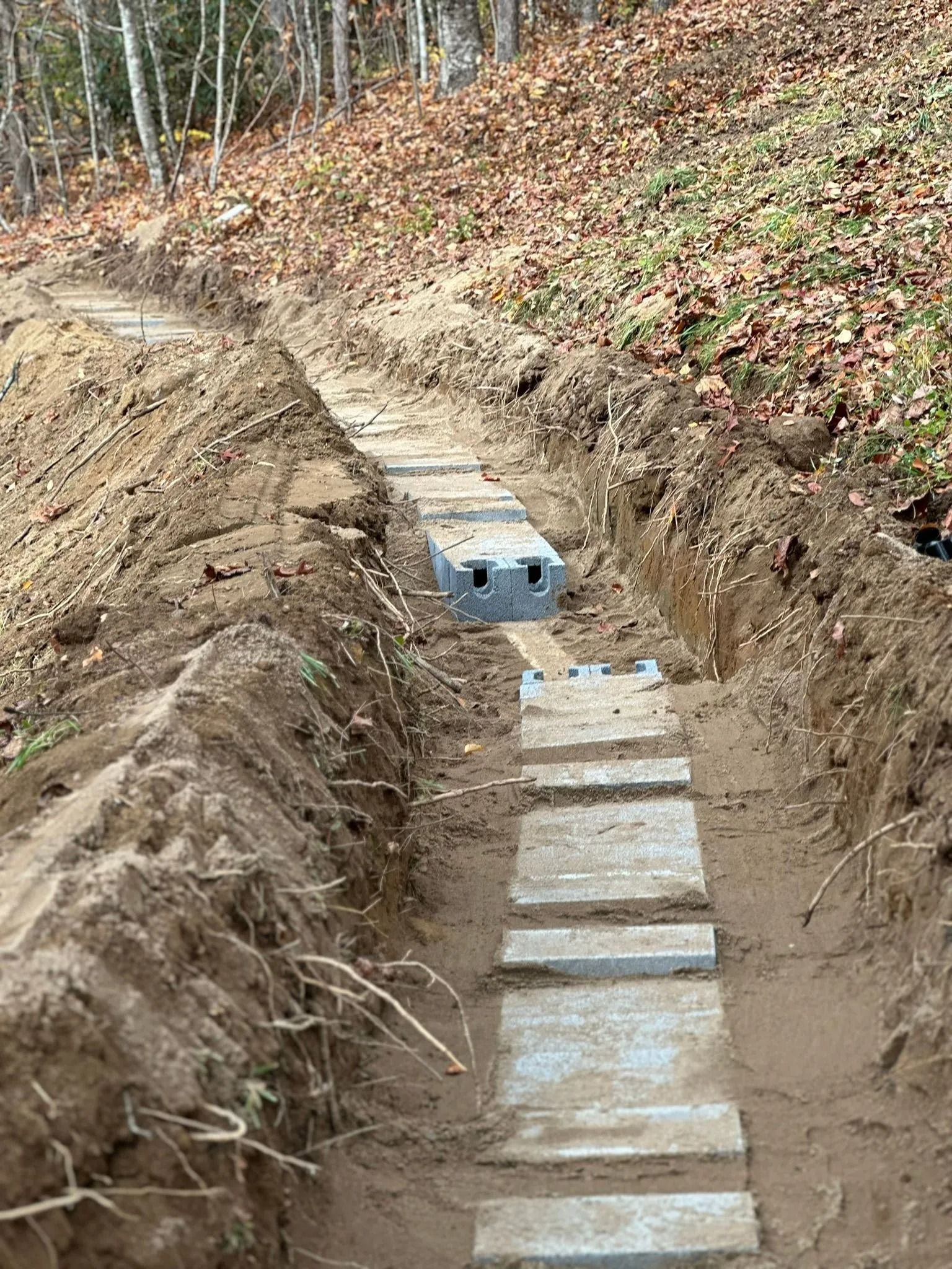 Trench with installed concrete steps and two gray concrete boxes. Dirt bank backdrop with some foliage.