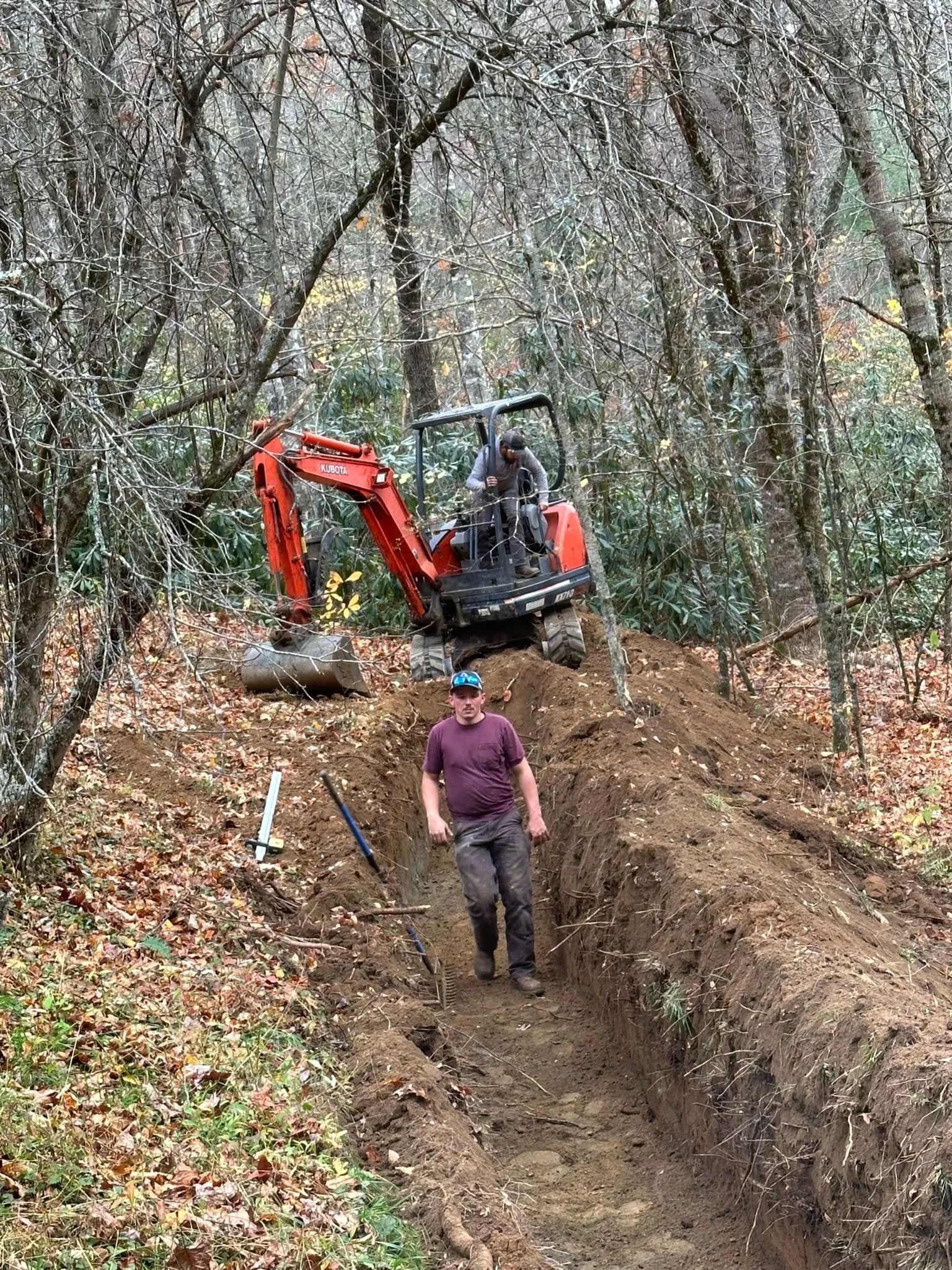 Man walks out of a trench. An excavator digs nearby in a forest.