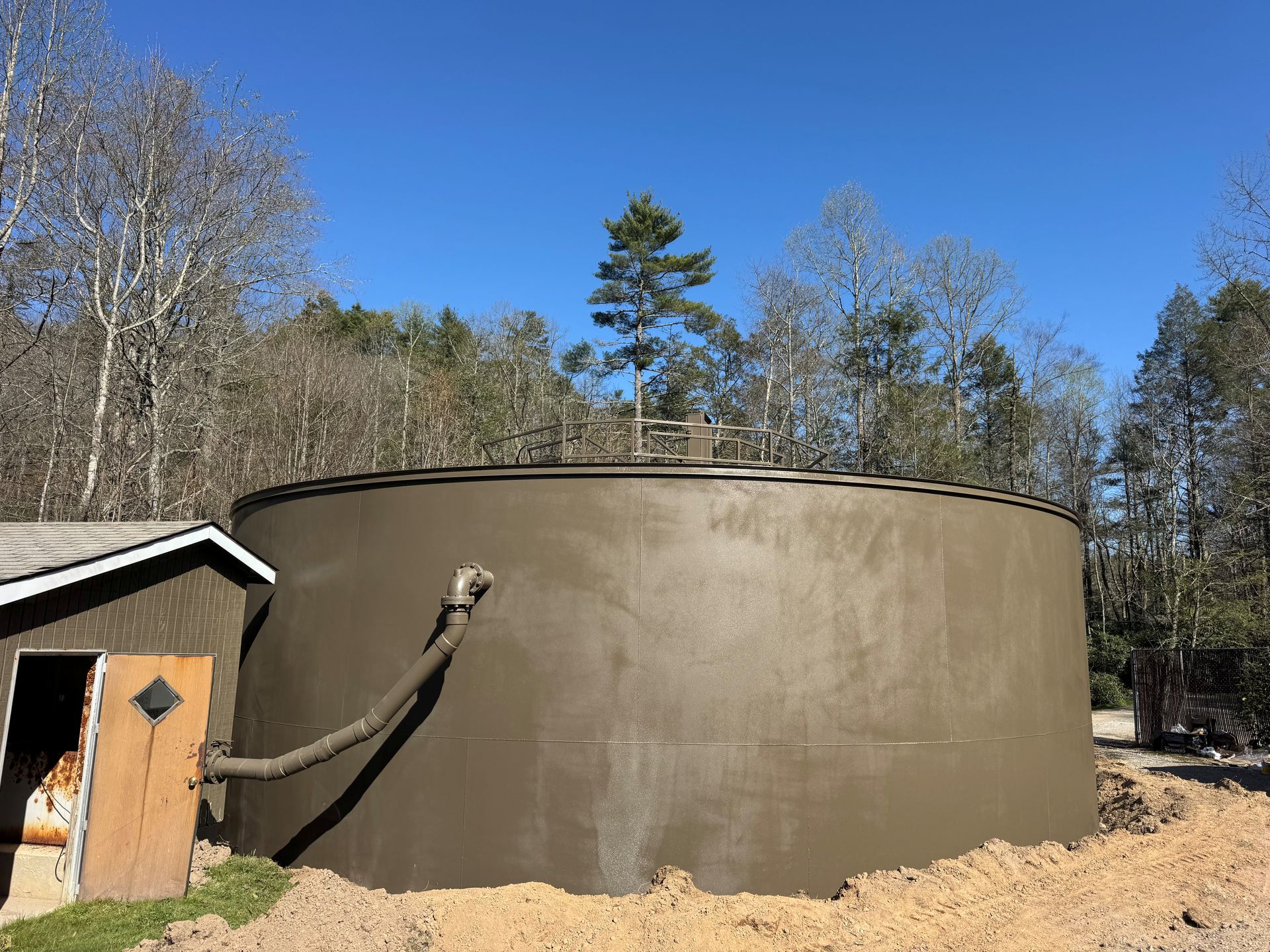 Large brown cylindrical water tank beside a small shed in a wooded area under a blue sky