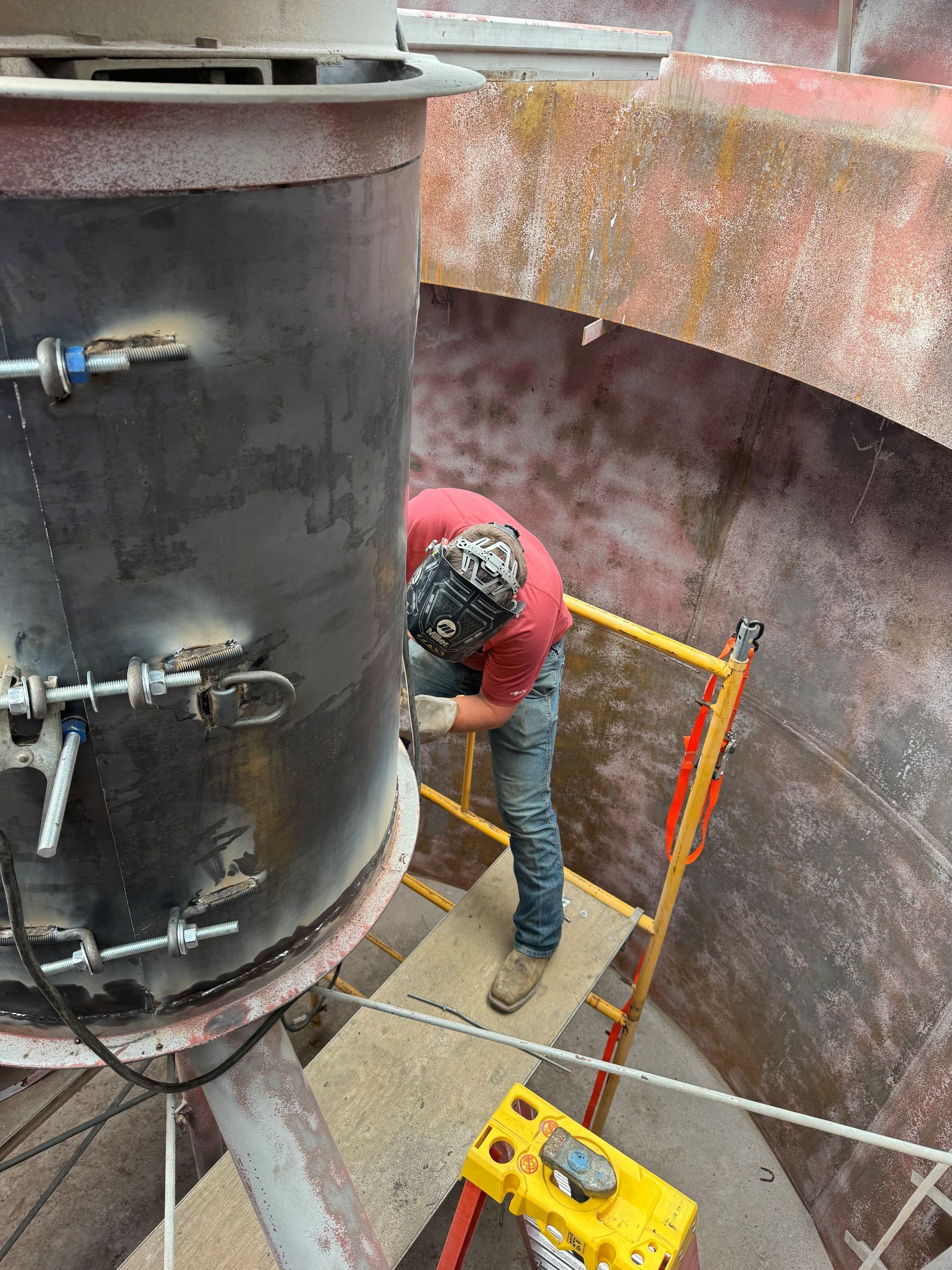 Worker in red jacket crouches inside a metal tank on a scaffold, with tools and piping nearby.