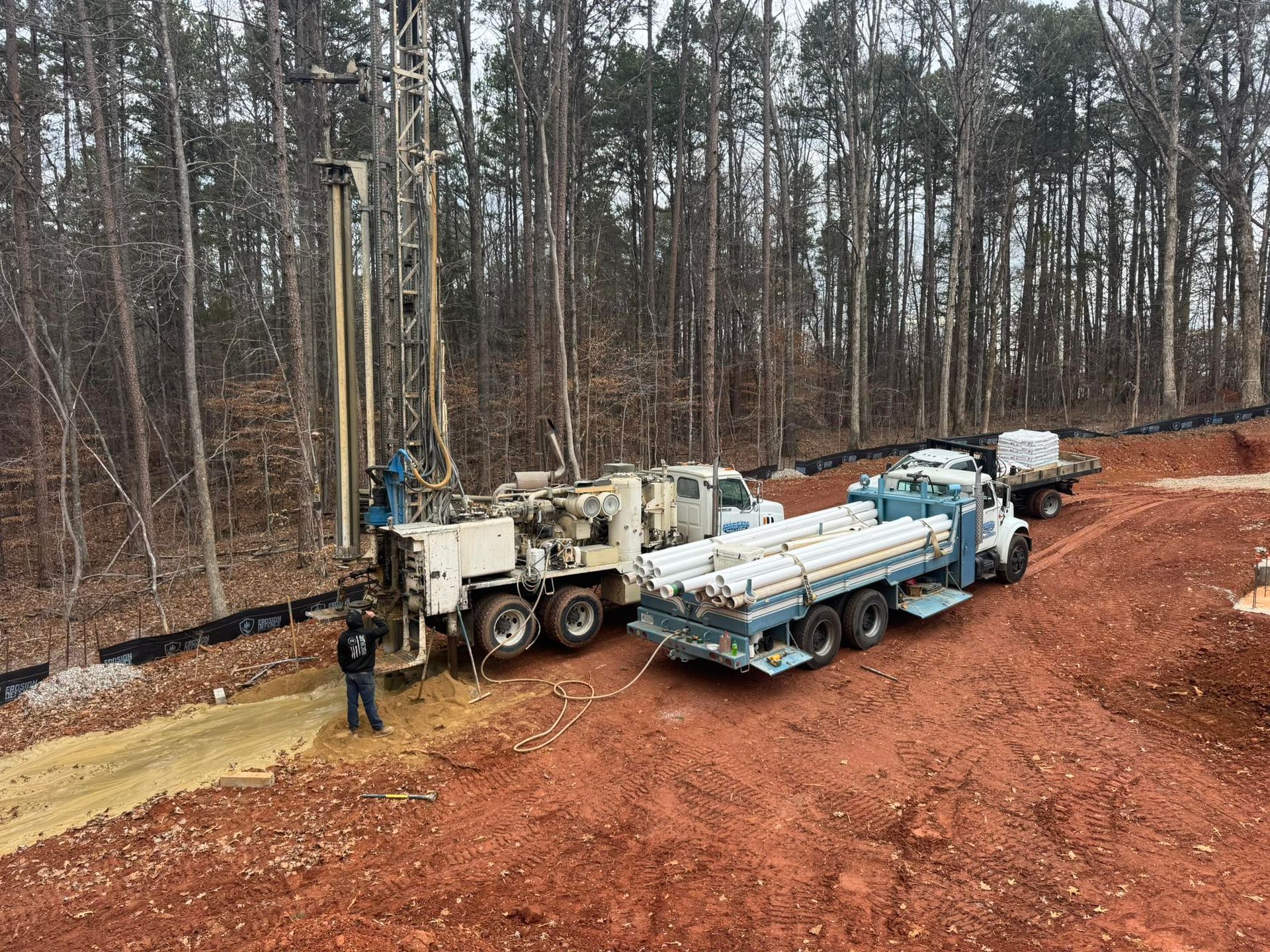 A large white drill rig parked on red clay soil near a forest, with a truck carrying steel drilling pipes attached.