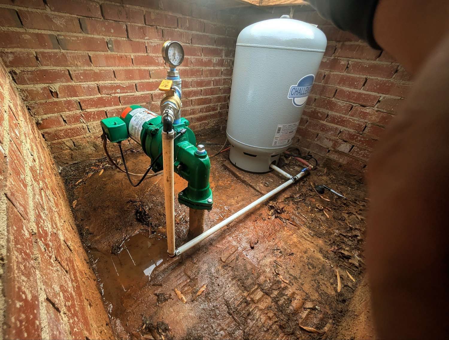 A green water pump and grey pressure tank situated in a damp, brick-walled enclosure on a dirt floor.
