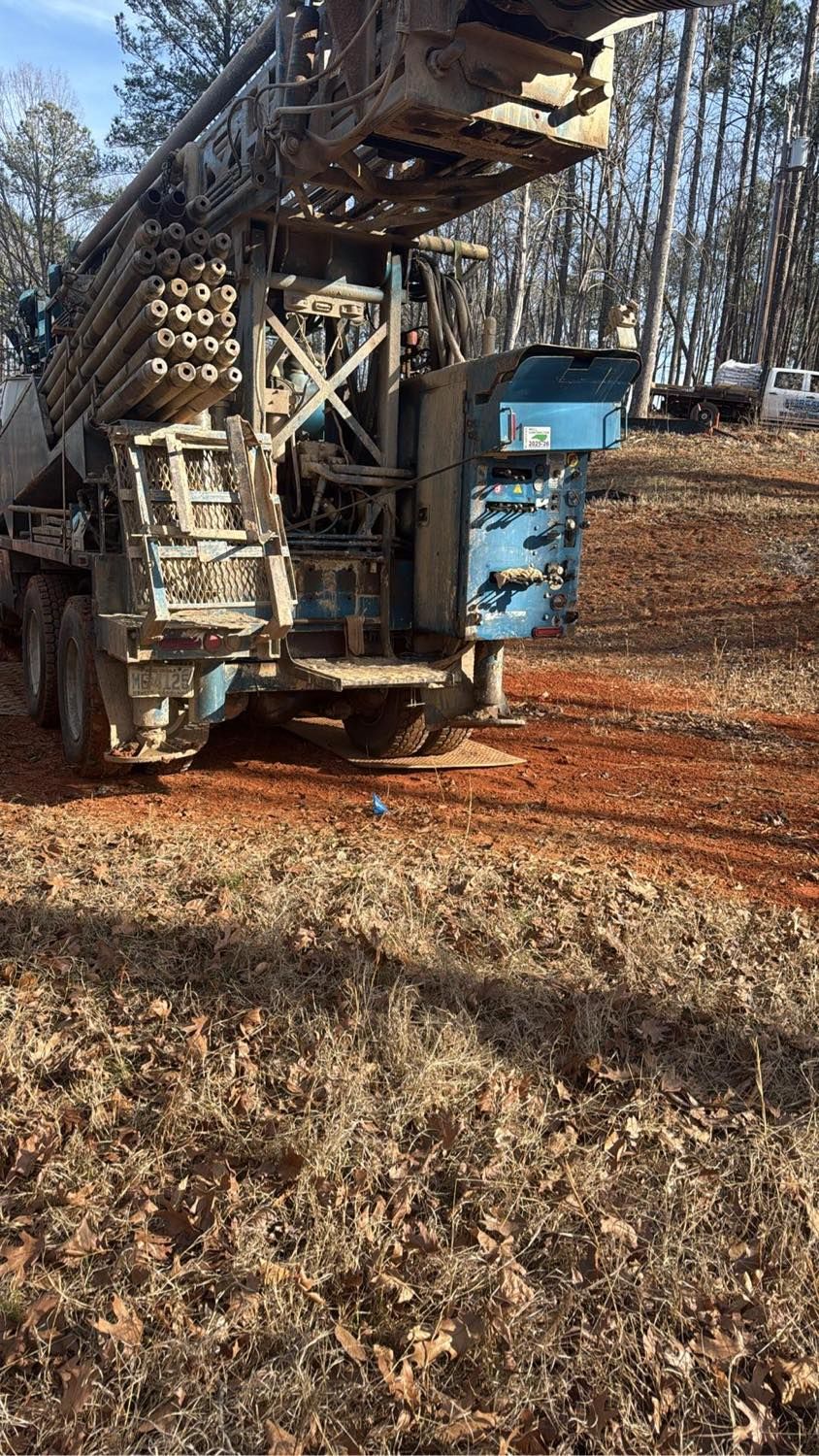 A truck-mounted drilling rig stands on a red dirt clearing near trees in a forested area.