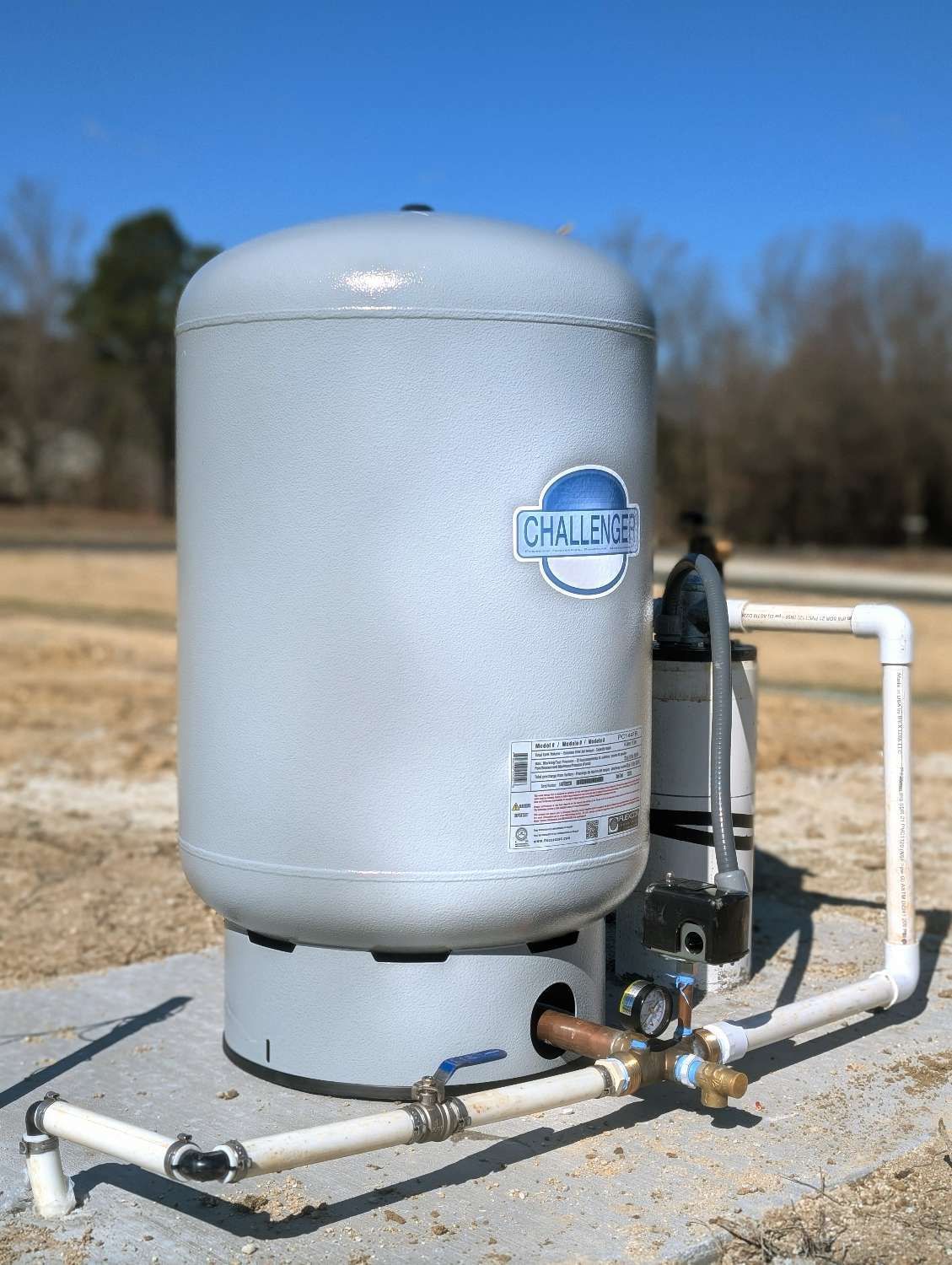 A large, light gray well pressure tank stands on a concrete pad with connected white PVC plumbing pipes and a gauge.