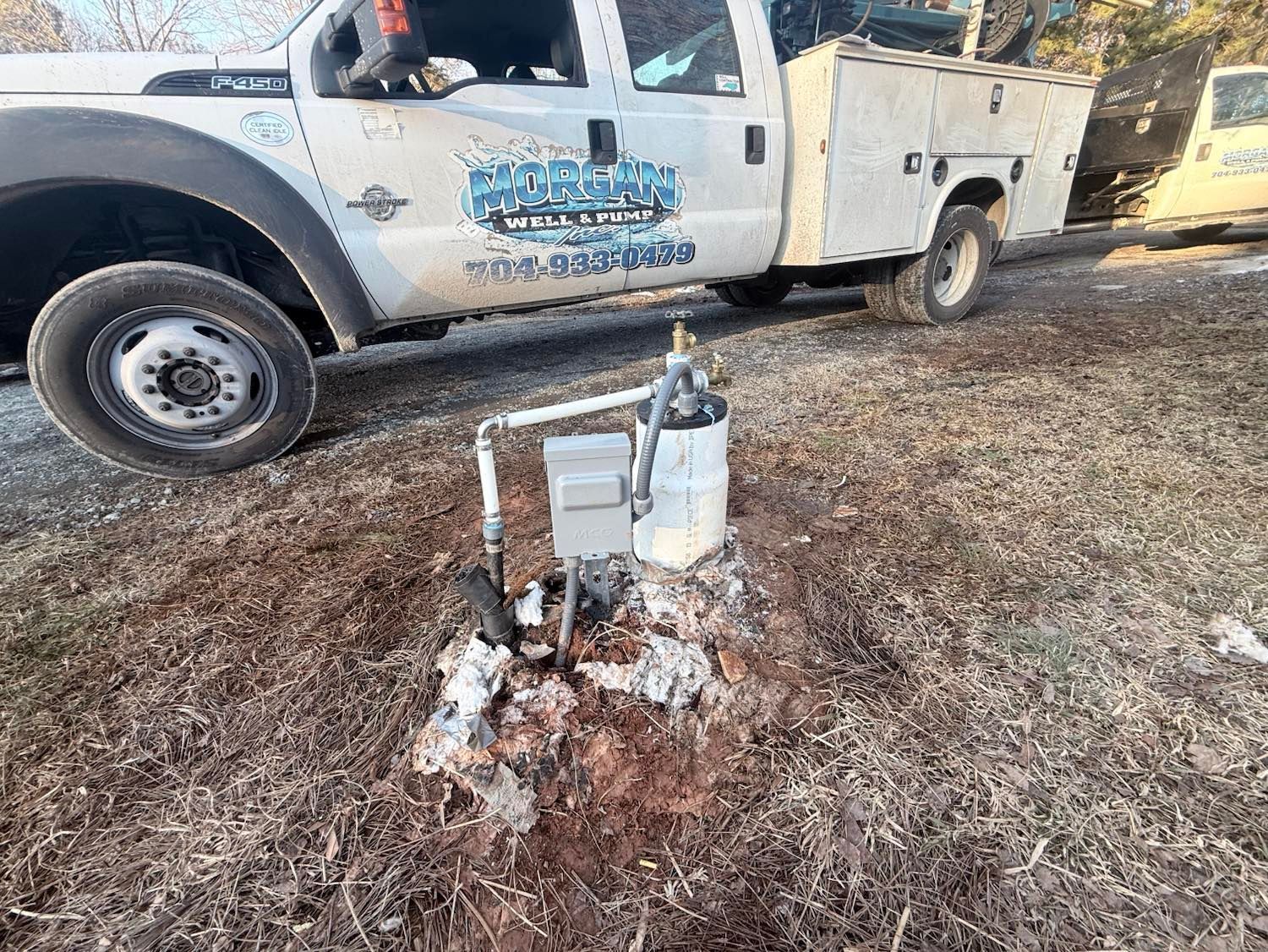 A white utility truck is parked on grass near a water well pump with an electrical box.