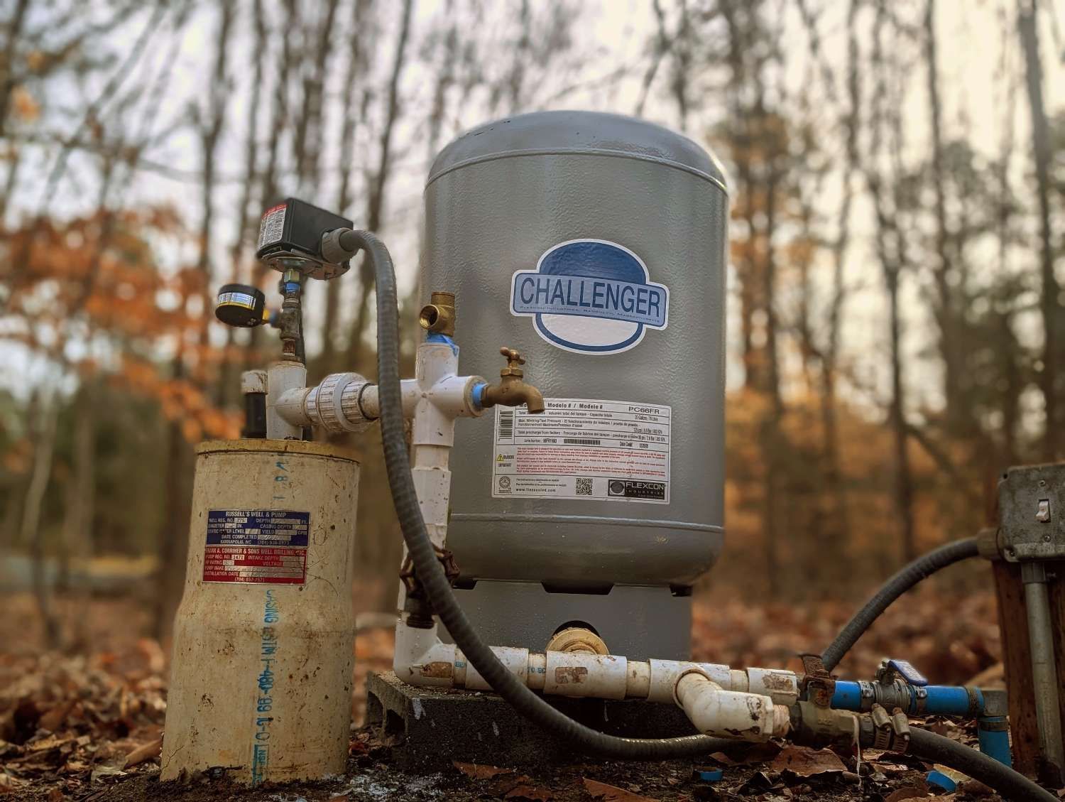 A Challenger well water pressure tank with attached pipes, gauges, and a filter, sitting outdoors on a wooded ground.