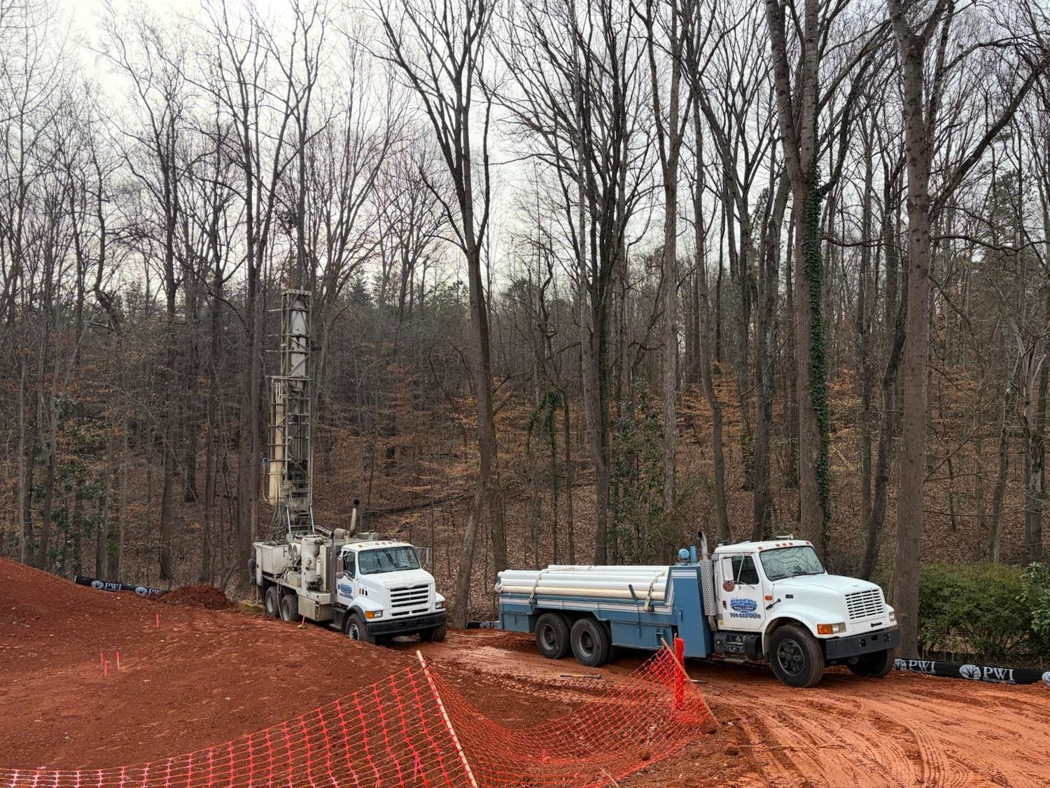 Two white industrial drill trucks parked on red dirt at a wooded construction site with orange plastic fencing.