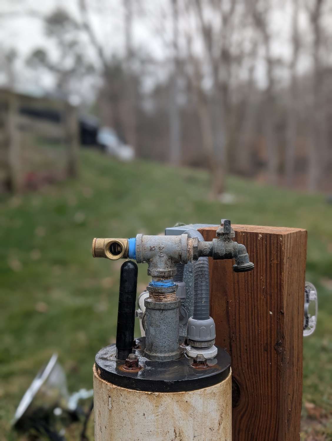 A metal wellhead pipe assembly with a spigot and valve, mounted on a wooden post outdoors against a blurred yard background.