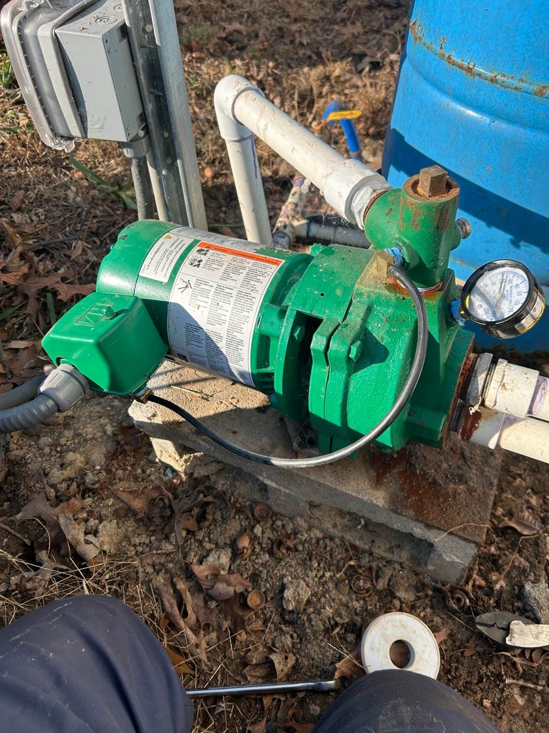A green water pump installed on a concrete block outdoors next to a blue tank and electrical conduit.