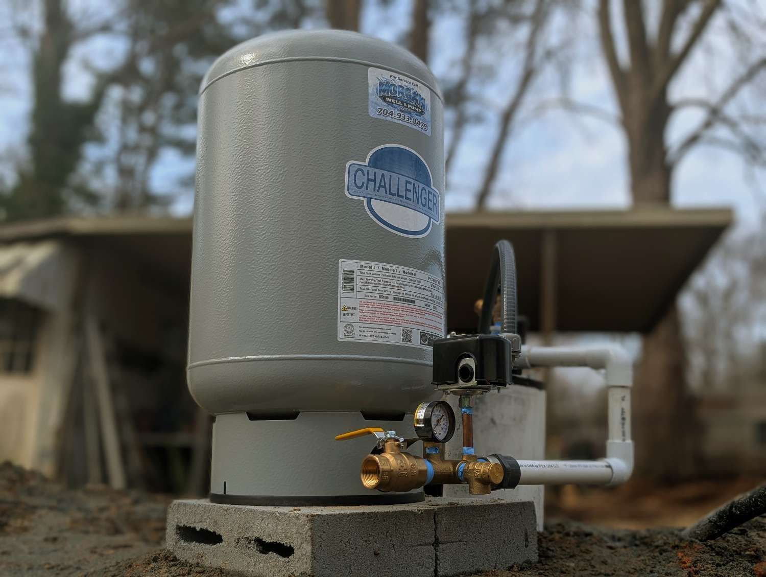 A gray Challenger brand pressure tank sits on a concrete block outdoors with plumbing, a pressure gauge, and a switch.