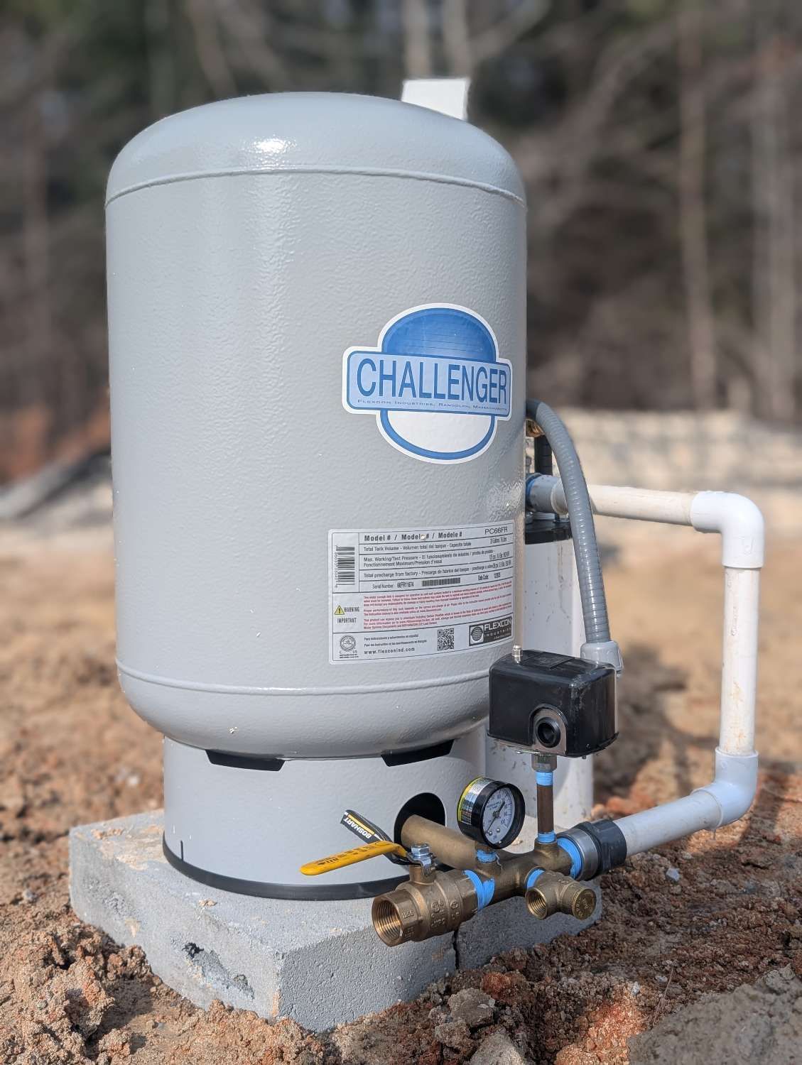 A gray Challenger pressurized water tank with a pressure gauge and plumbing connections sitting on a concrete block.