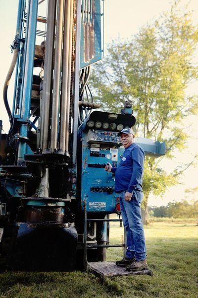 A person wearing a blue shirt and jeans stands at the controls of a blue industrial drill rig in a field.