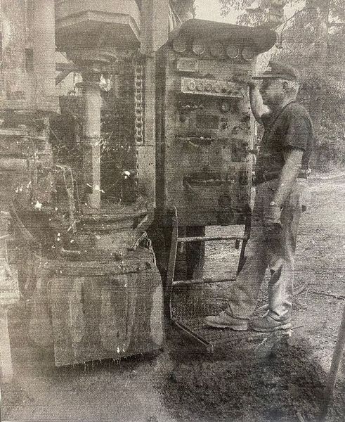 A person stands at a control panel operating a vertical drilling rig outdoors.