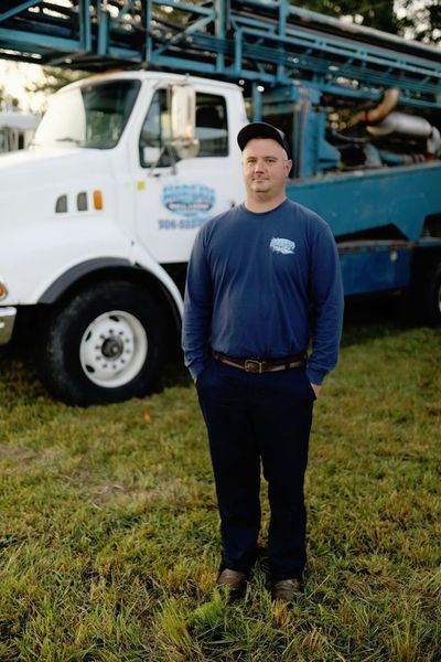 A person stands in a grassy field in front of a white and blue work truck on a sunny day.