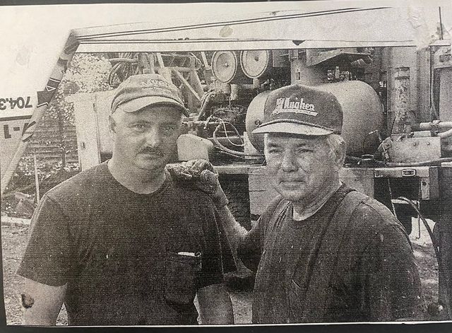 Two men in baseball caps and dark t-shirts stand in front of industrial machinery in a black and white photo.