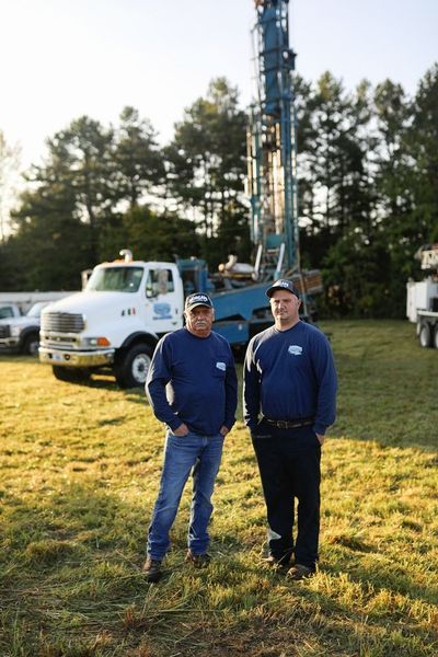 Two people wearing blue long-sleeve shirts stand in a grassy field in front of a white well-drilling truck.
