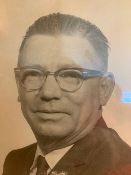 A portrait of a person with short hair, wearing glasses, a white collared shirt, and a dark jacket.