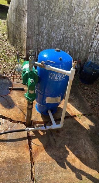 A blue Water Works pressure tank and green pump connected by white PVC pipes on a concrete base outdoors.