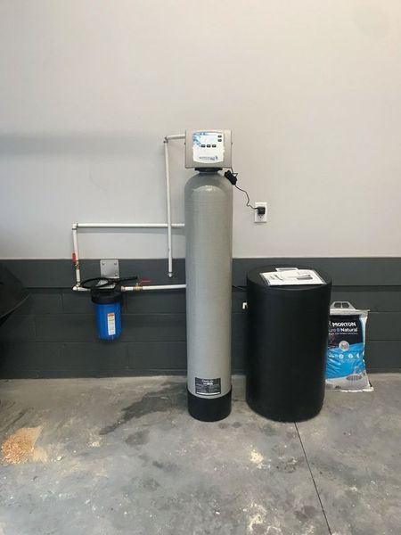 A home water softener system with a tall gray tank, black brine tank, and blue filter housing mounted on a garage wall.