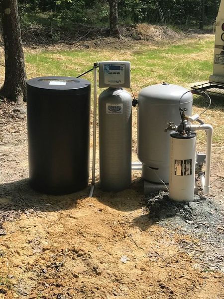 A water treatment system with a black brine tank, a grey softener, a pressure tank, and a filter, set on a dirt ground.