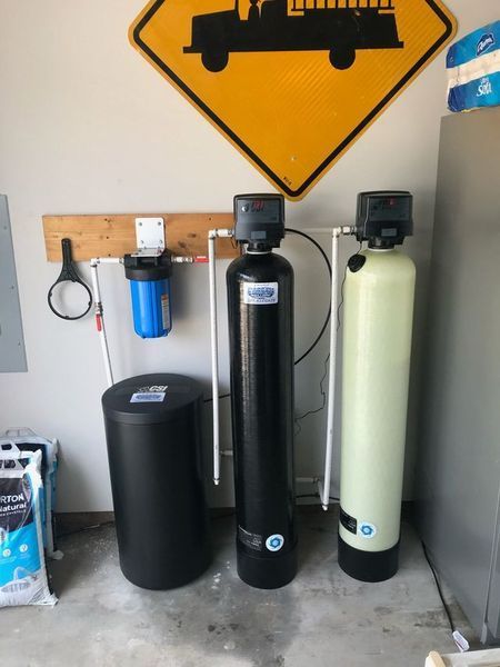 A water filtration system in a garage featuring a blue sediment filter, a black brine tank, and two tall water tanks.