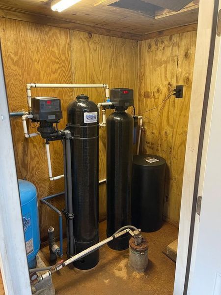 A water filtration and softener system with two black tanks and a blue pressure tank in a plywood-walled utility closet.