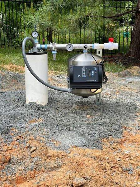 A well pump system with a metal pressure tank and gauge sits on a gravel patch in a yard near a black fence.