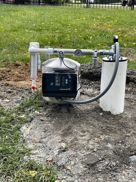 A metal water pump system with a digital control box and pressure gauge, installed on a dirt patch near a grassy lawn.