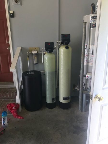 A home water filtration and softening system with two tall tanks and a brine tank in a garage next to a water heater.