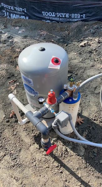 A grey water pressure tank and filter system sit on the ground next to pipes and valves in a dirt-filled construction area.