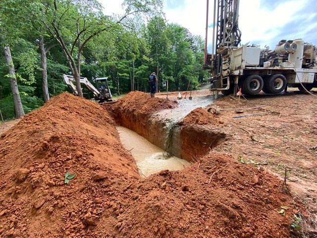 A drilling rig operates next to a deep trench filled with muddy water on a construction site with trees in the background.