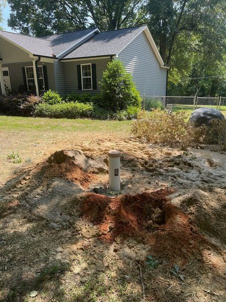 A vertical white pipe stands in a mound of upturned dirt in a residential yard in front of a gray house.