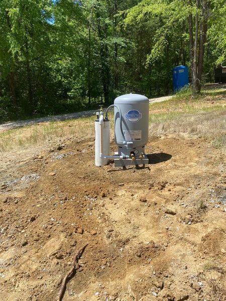 A gray water pressure tank and white PVC pipe stand on a patch of bare dirt in a wooded, outdoor area.