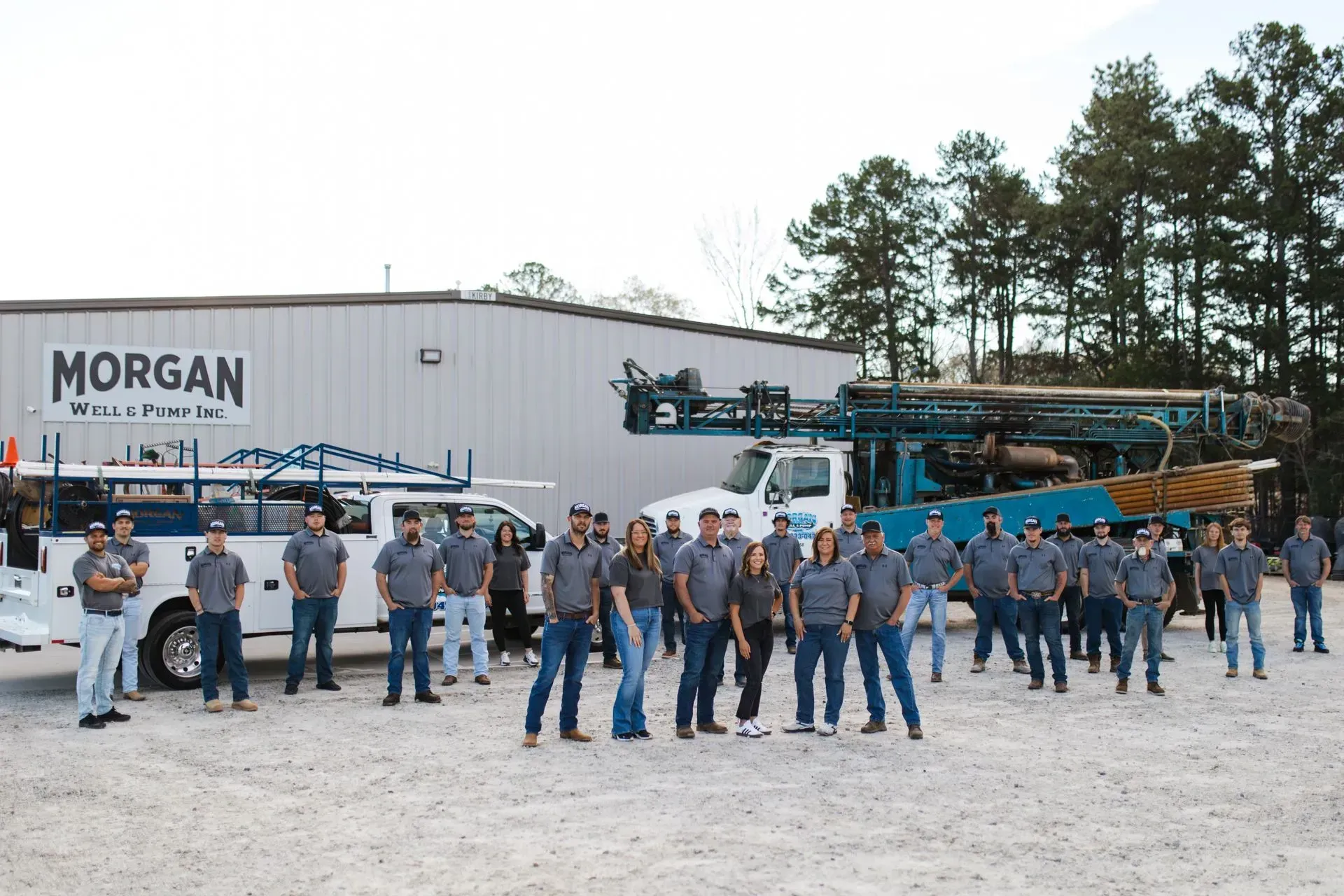 A large group of people in uniform posing in front of a white building with Morgan Water & Pump signage and a drill truck.