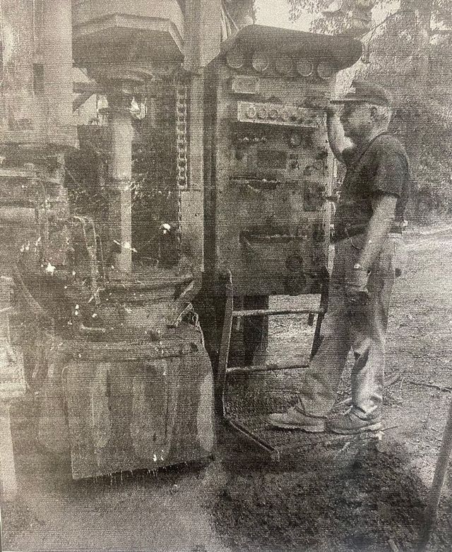 A person wearing a cap and work clothes stands on a platform operating the controls of a large industrial drilling rig.