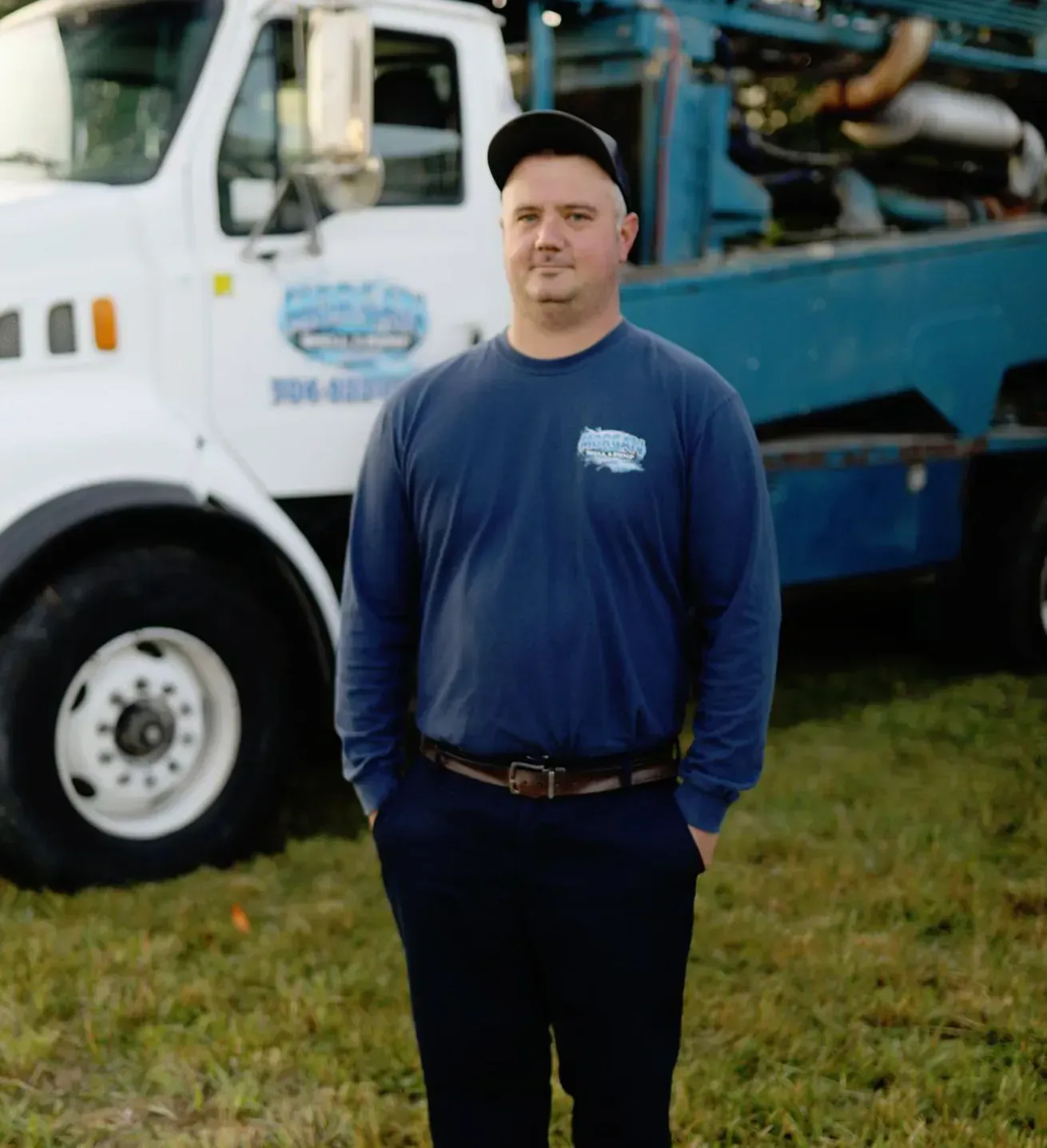 A person in a blue long-sleeve shirt and cap stands in a grassy field in front of a parked white and blue service truck.
