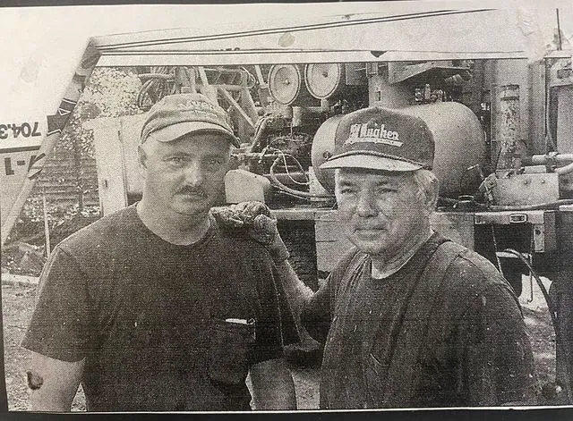 Two people wearing baseball caps stand outdoors in front of a piece of industrial machinery.