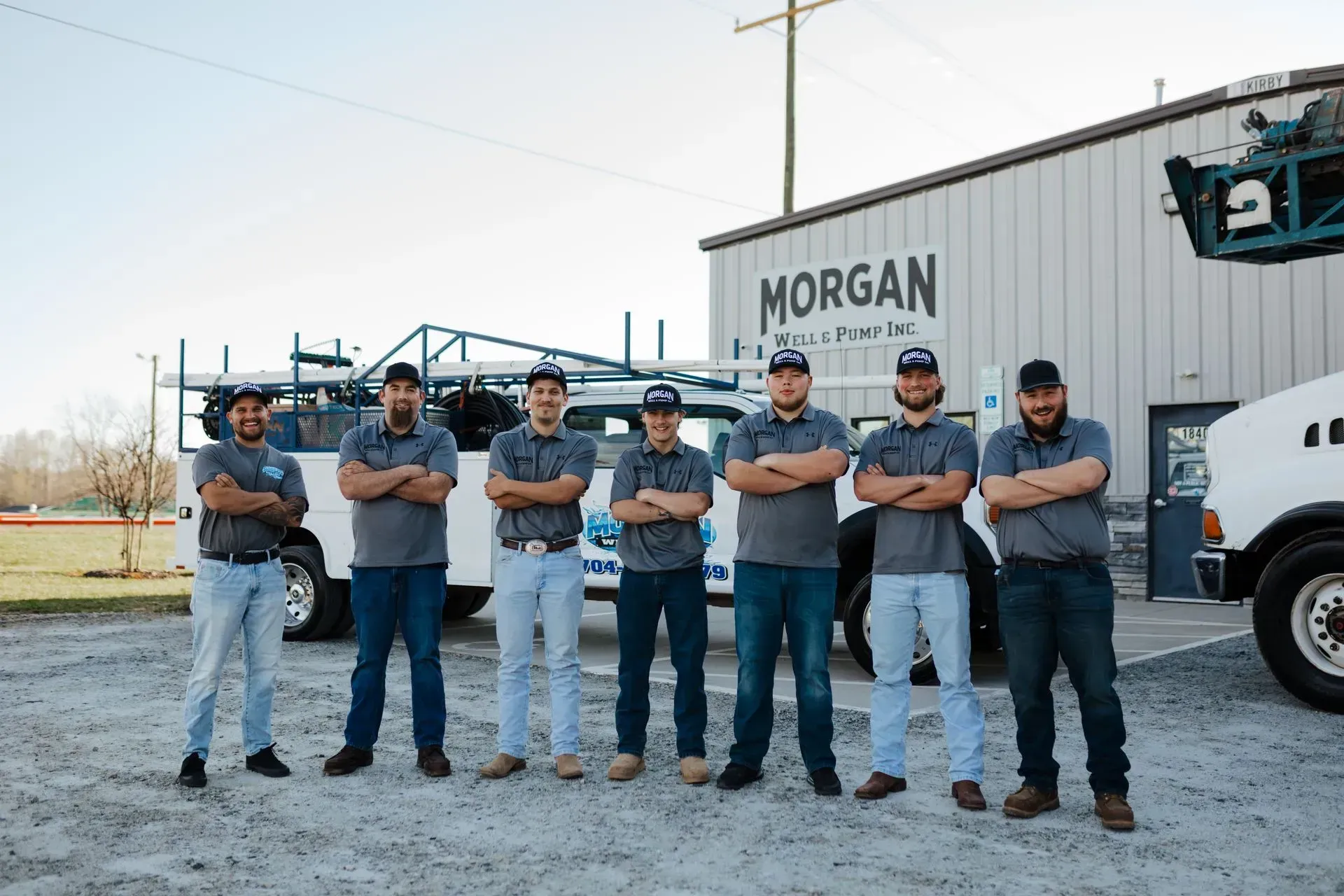 Seven employees in gray shirts and hats stand with arms crossed in front of a Morgan industrial building and work truck.