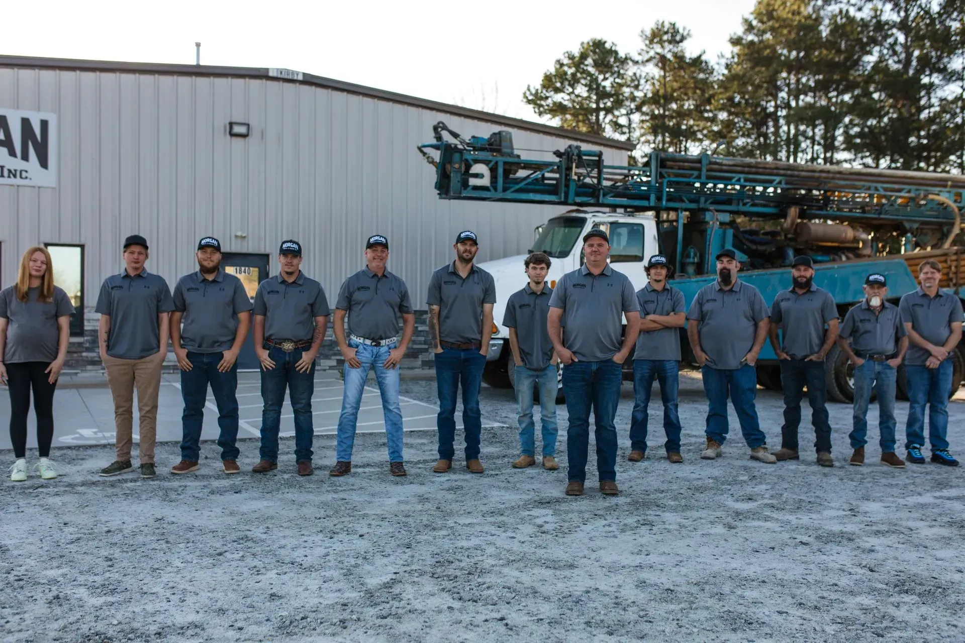 A group of employees in matching grey shirts standing in a line in front of a drill rig parked at an industrial facility.