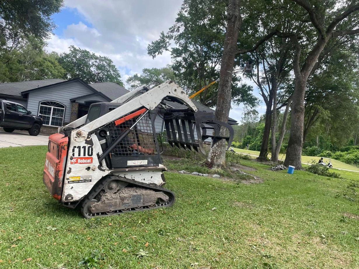 Compact loader on a grassy yard beside a house, with a boom attachment and trees in the background.