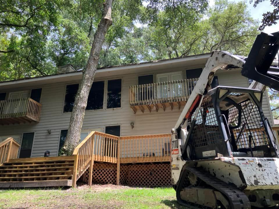House exterior with a wooden deck and steps, trees overhead, and a skid-steer loader parked in the yard.