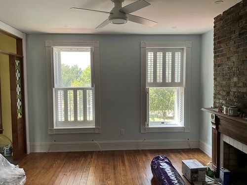 Empty living room with gray walls, hardwood floors, windows, ceiling fan, and brick fireplace.