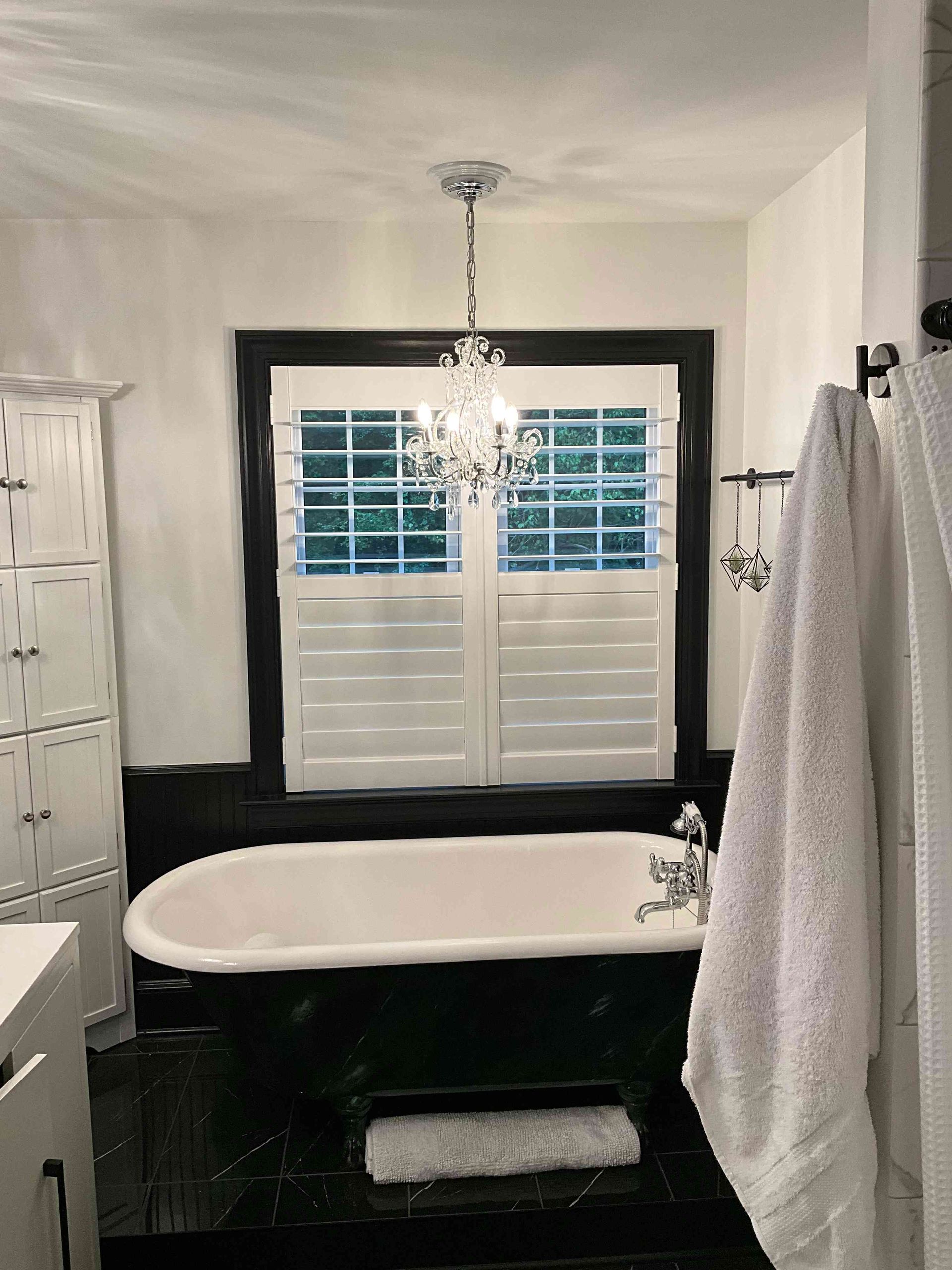 Black-and-white bathroom with freestanding tub, chandelier, shuttered window, and hanging towels