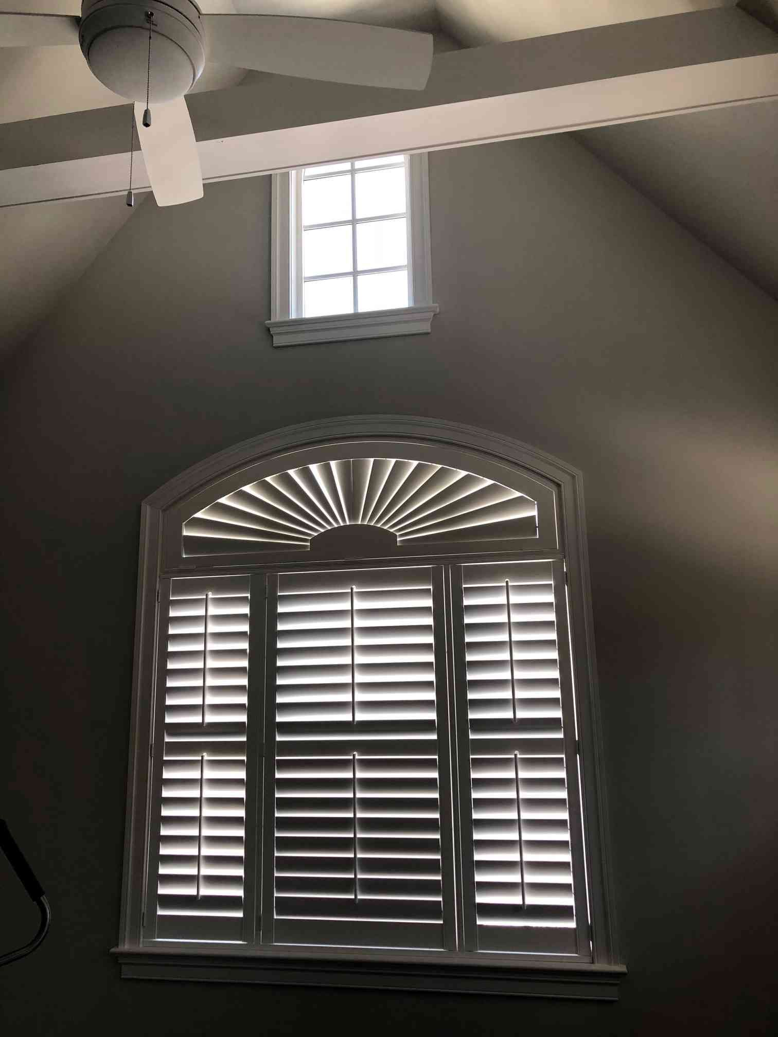 Gray bedroom wall with arched shutters and a small window above, beneath a ceiling fan in a vaulted ceiling