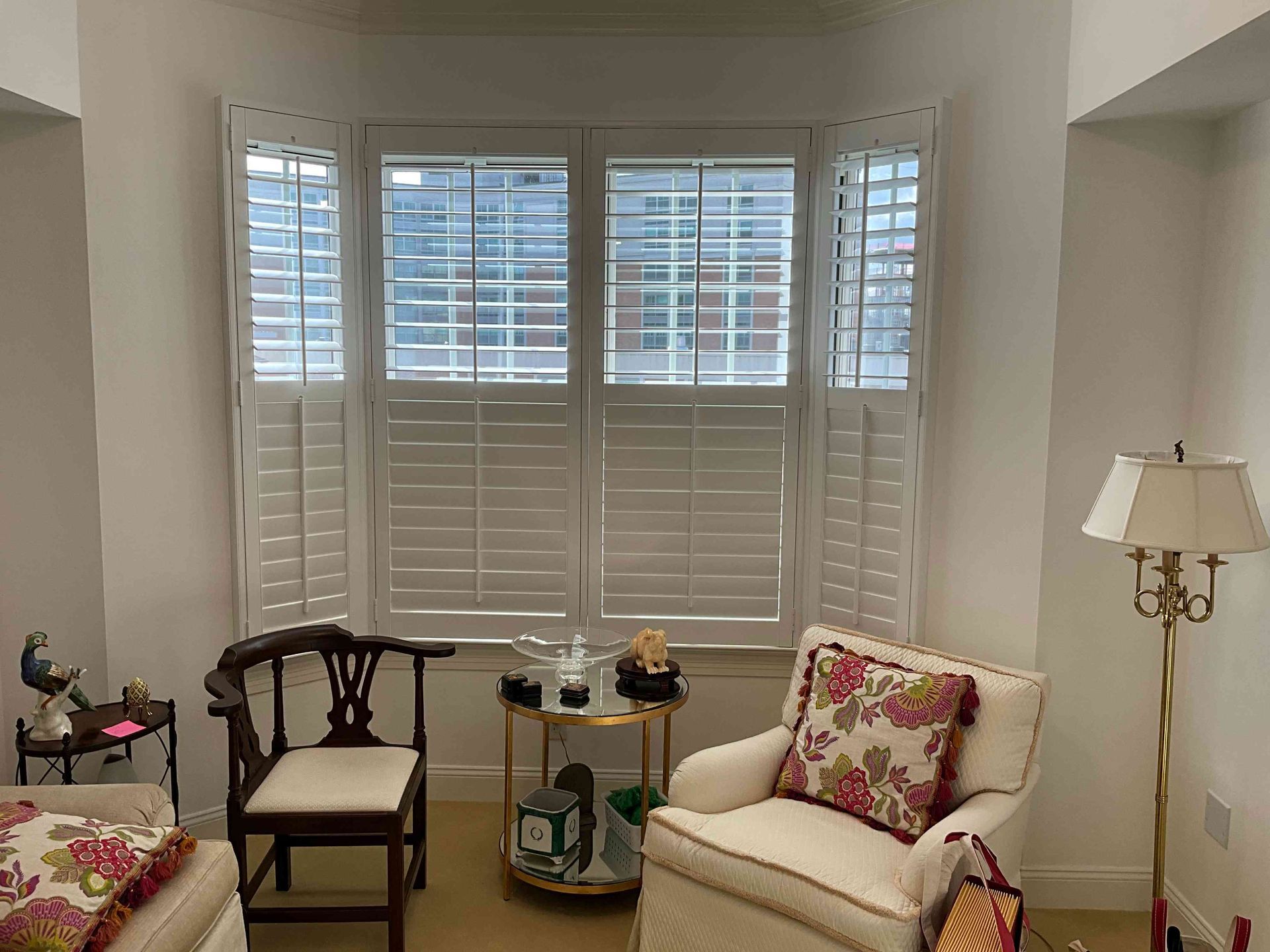 Bright sitting room with white shutters, floral chairs, and a small side table near a lamp.