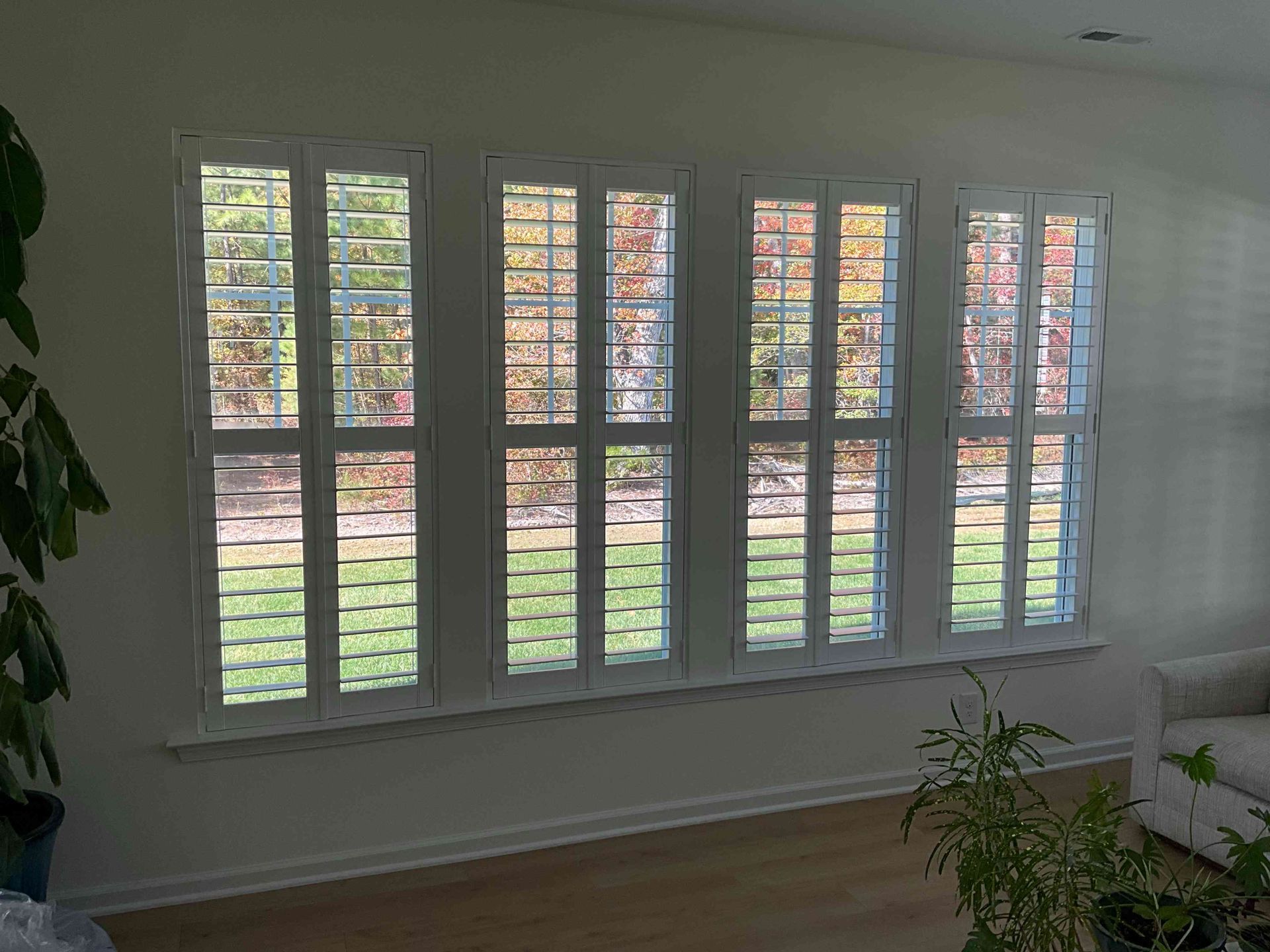 Bright living room with large windows, white shutters, and a view of the green backyard.