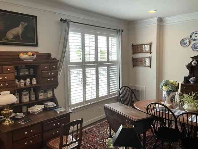 Bright dining room with wooden table, hutch, window shutters, and decorative plates on the wall.