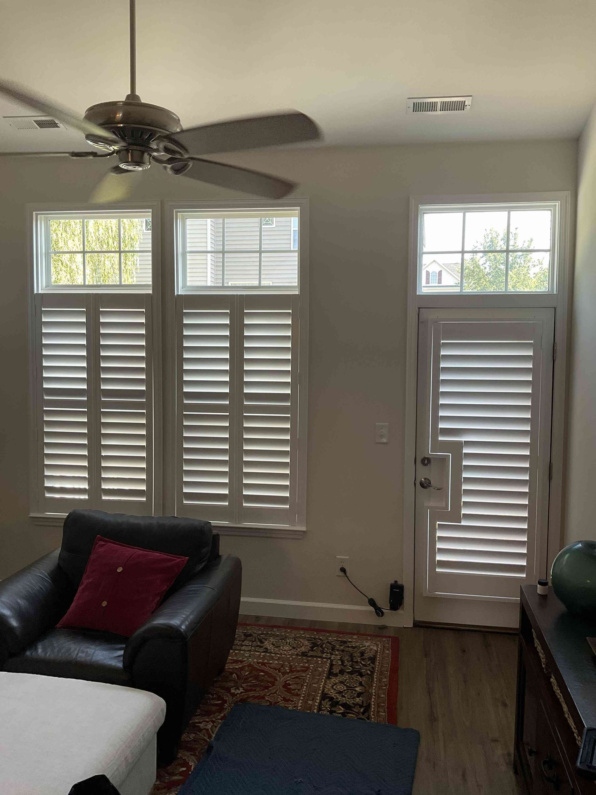 Living room with black armchair, red pillow, shutters, front door, and ceiling fan.